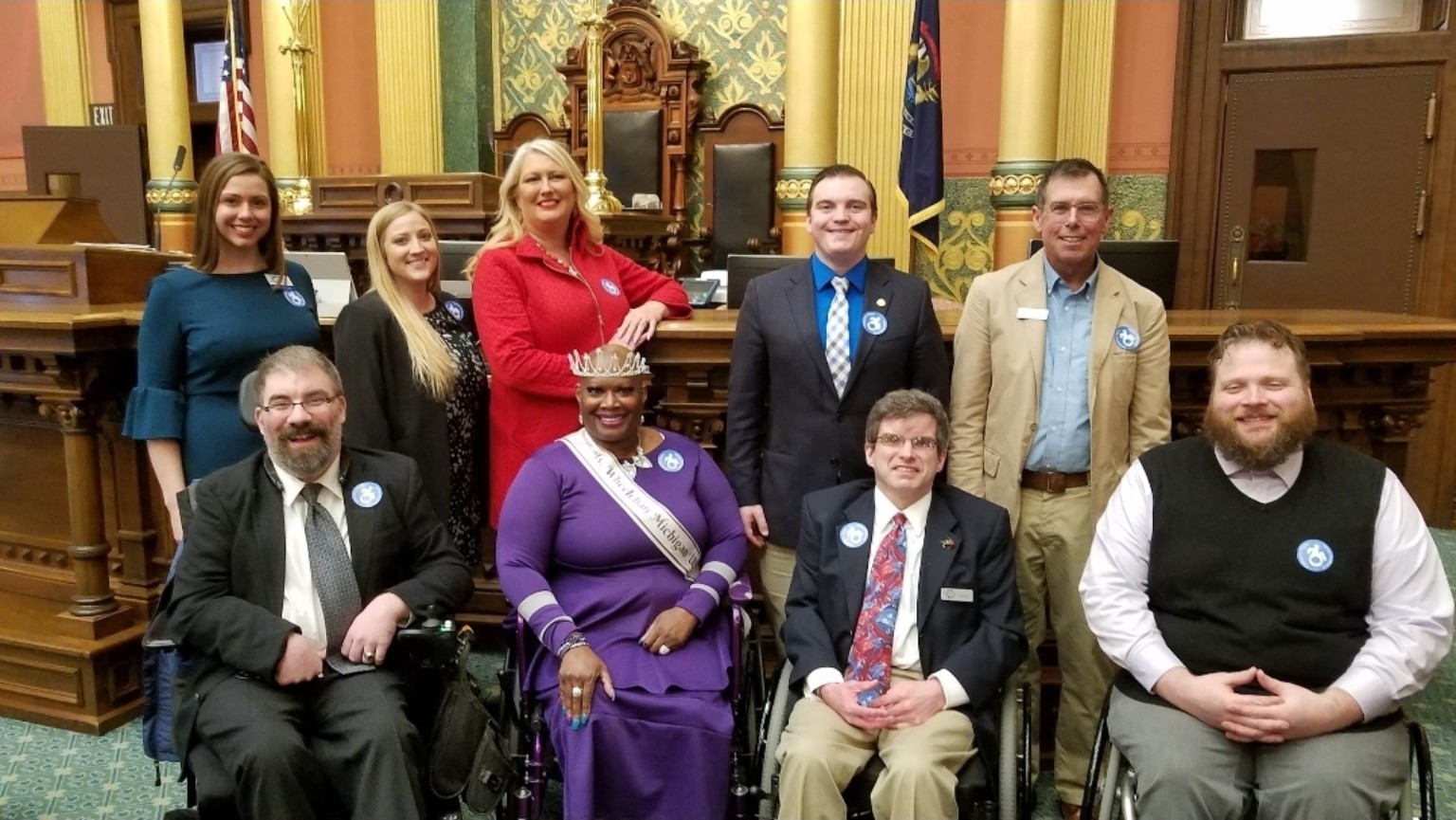 Dr. Kimberly Y. Barrington, wearing purple and seated in her wheelchair, poses with a group of advocates in a legislative chamber. Several wear blue accessibility-symbol buttons.