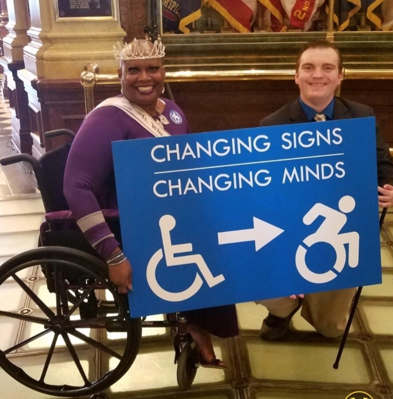 Dr. Kimberly Y. Barrington, wearing her crown and seated in her wheelchair, and Beau LaFave, kneeling beside her, hold the large blue Changing Signs, Changing Minds sign.