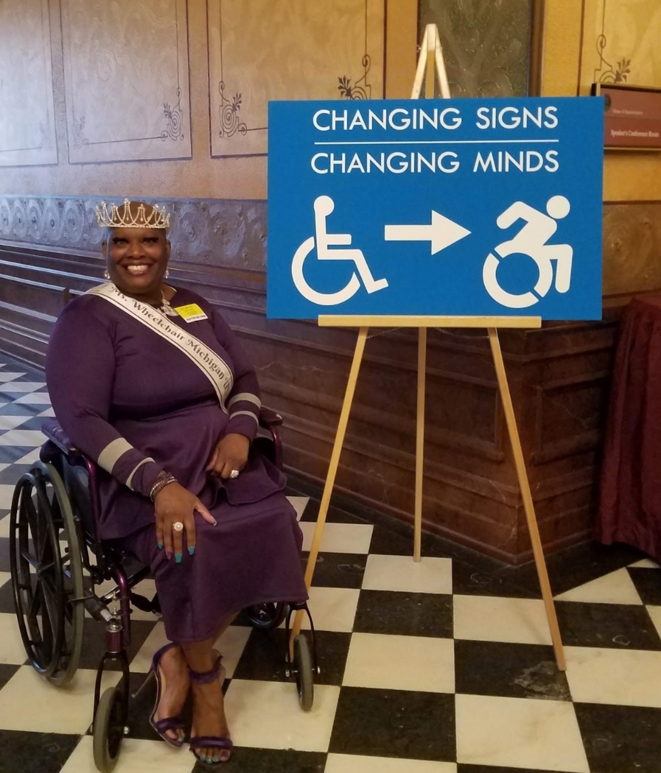 Dr. Kimberly Y. Barrington, wearing a crown and purple dress, sits in her wheelchair beside an easel displaying the large blue Changing Signs, Changing Minds sign.