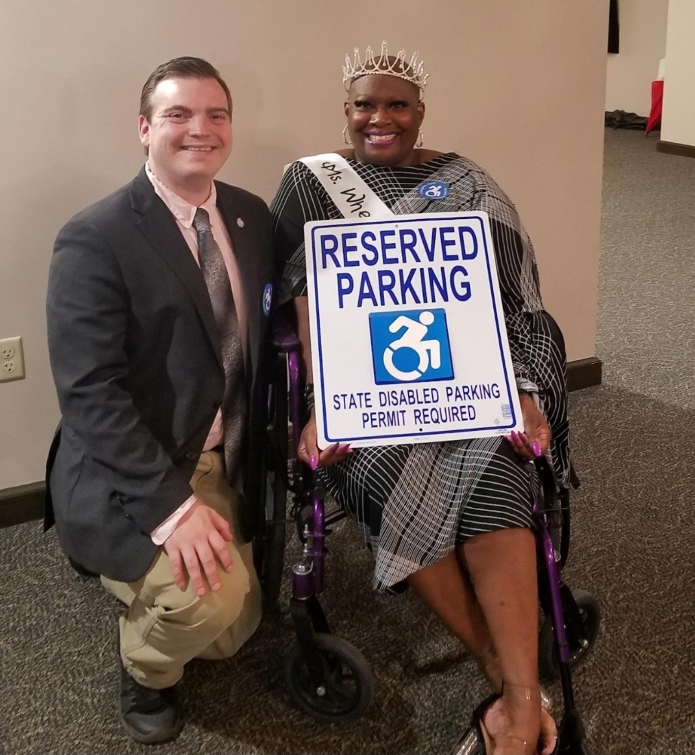 Dr. Kimberly Y. Barrington, seated in her wheelchair, and Beau LaFave, kneeling beside her, hold a reserved-parking sign featuring the forward-moving accessibility symbol. The sign reads State Disabled Parking Permit Required.
