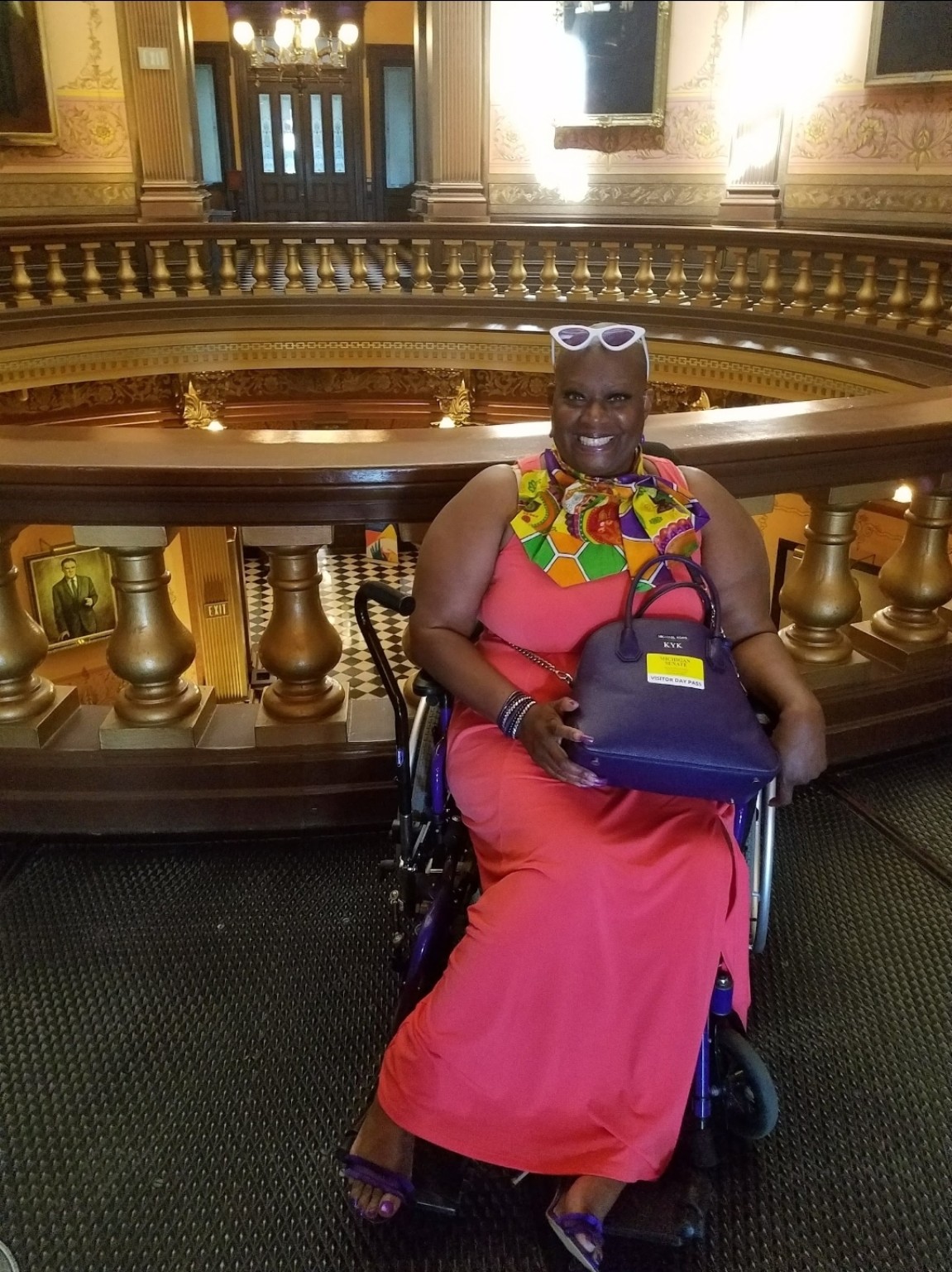 Dr. Kimberly Y. Barrington smiles from her wheelchair beside the ornate balcony railing inside the Michigan Capitol, wearing a coral dress, colorful scarf, and white sunglasses on her head.