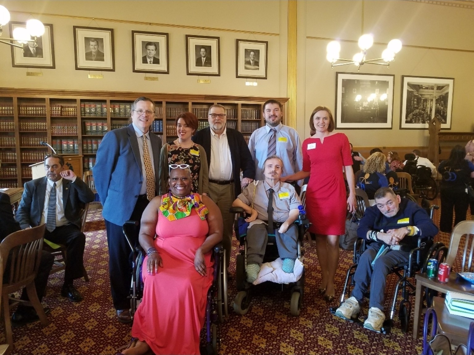 Dr. Kimberly Y. Barrington, seated in her wheelchair and wearing a coral dress, poses with advocates and legislators in a Capitol room lined with bookshelves and framed portraits.