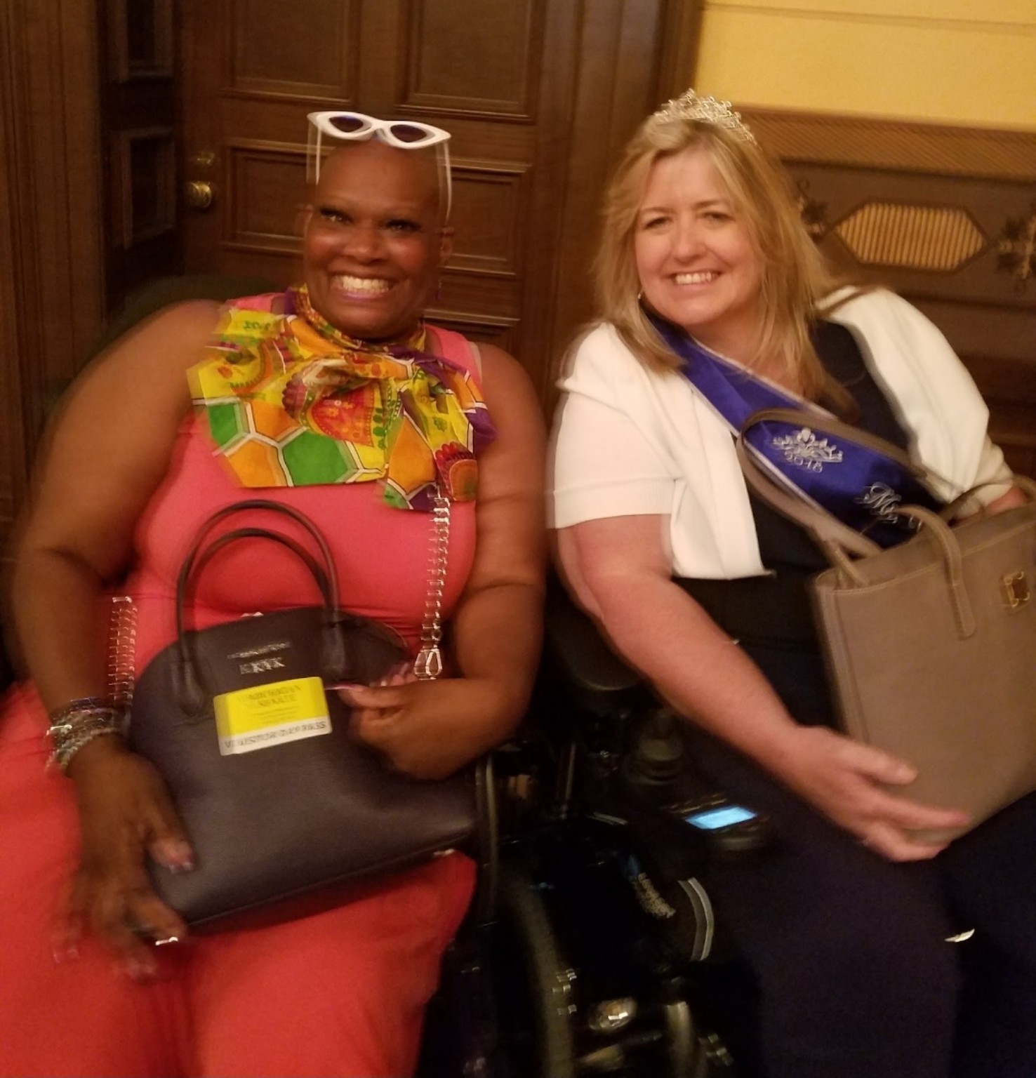 Dr. Kimberly Y. Barrington and Ms. Wheelchair America smile together, seated in their wheelchairs. Ms. Wheelchair America wears a crown, white jacket, and purple sash.