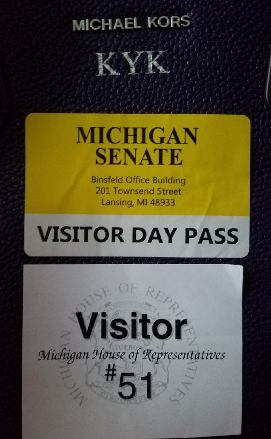 Two visitor passes on a purple handbag: a yellow and white Michigan Senate visitor day pass and a white Michigan House of Representatives visitor pass numbered 51.