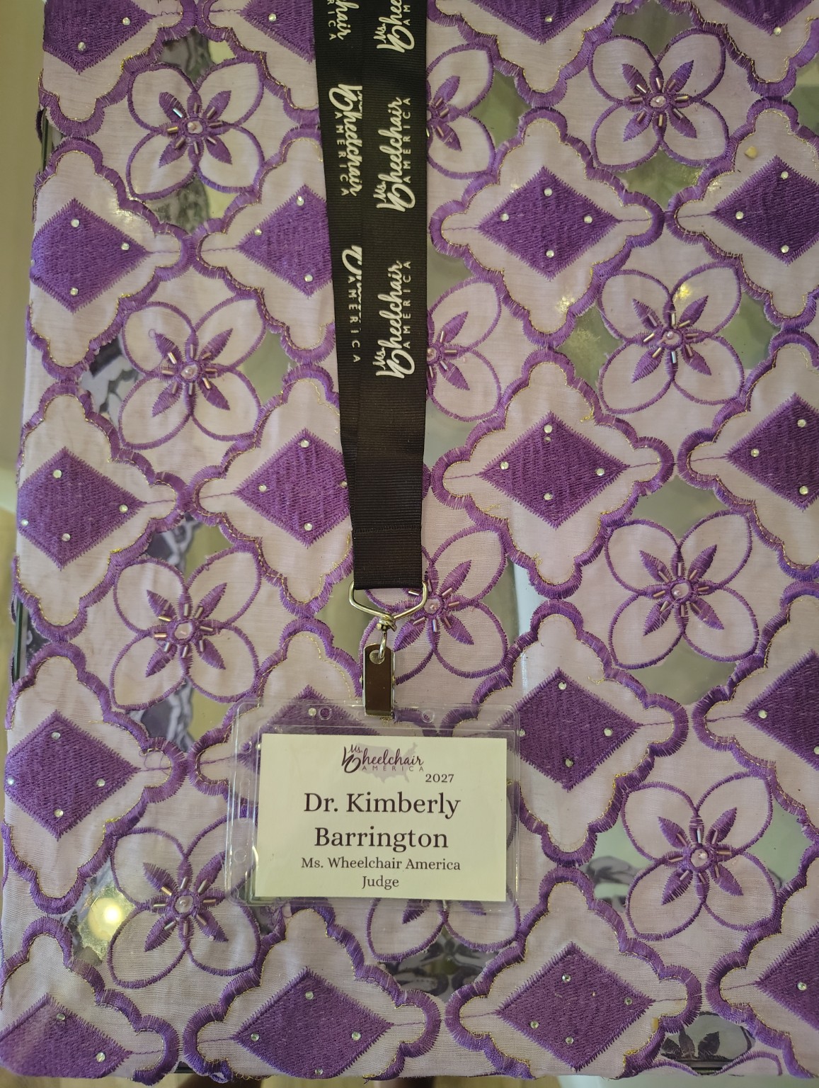Dr. Kimberly Barrington’s Ms. Wheelchair America 2027 judge badge and black lanyard resting on purple floral fabric.