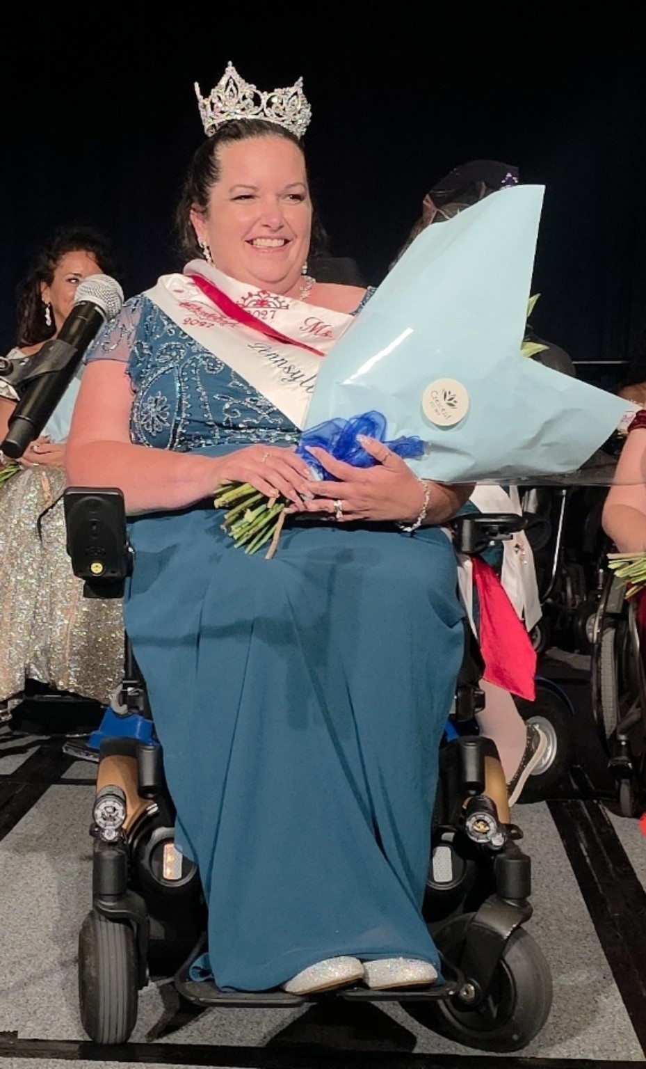 The newly crowned Ms. Wheelchair America 2027 smiles in a turquoise gown, wearing her crown and sash and holding a bouquet of flowers.