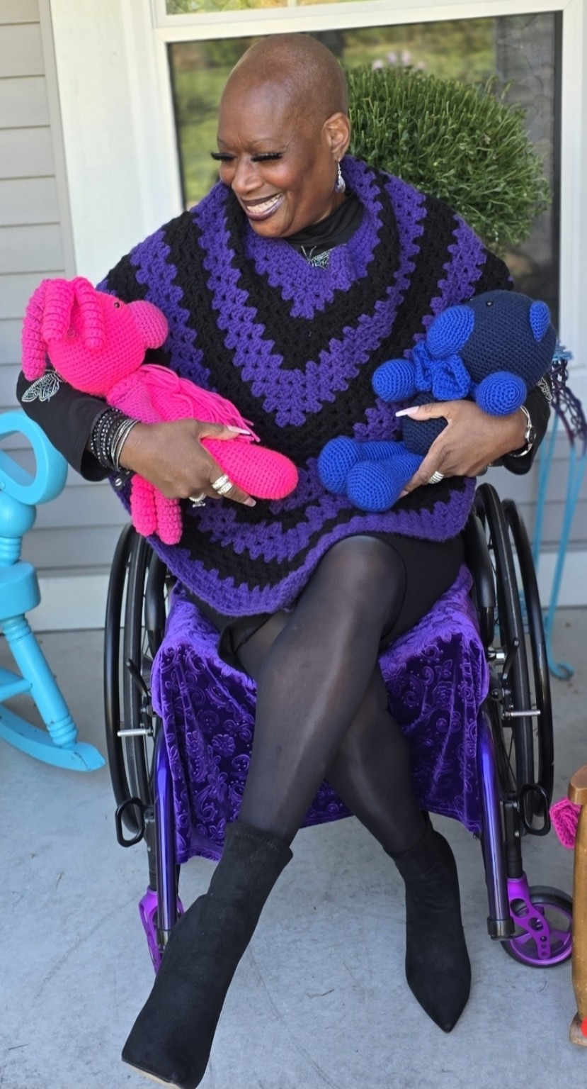 Dr. Kimberly smiles at the pink crocheted bear in one arm while holding a blue crocheted bear in the other, wearing Shana’s poncho.