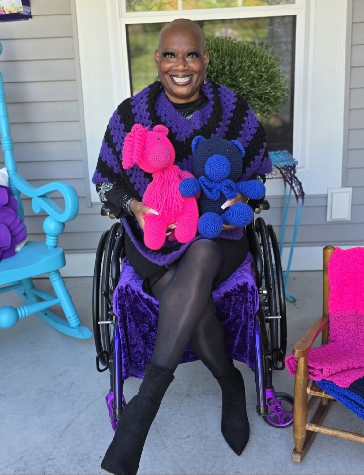 Dr. Kimberly wears Shana’s purple and black poncho and holds two colorful crocheted bears, one bright pink and one blue, while seated in her wheelchair.