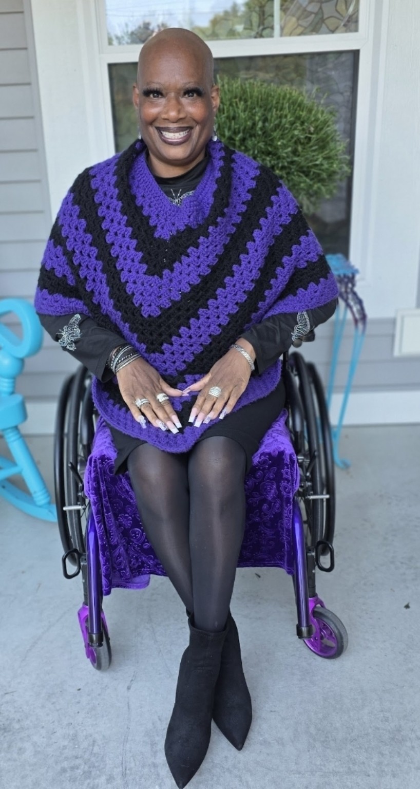 Dr. Kimberly smiles from her wheelchair while wearing a purple and black crocheted poncho made by Shana Thomas.